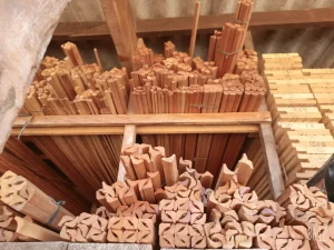 Área de armazenamento com peças de madeira dispostas verticalmente, incluindo molduras simples e perfis detalhados. À direita, diversas tábuas exibem marcações manuscritas em azul, como “Q18”, “Q16”, “Q14”, “Q12”, “Q10”, “Q8”, “Q6”, “Q4”, “Q2” e “Q1”. O cenário sugere organização para carpintaria ou controle de inventário.