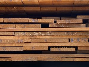 Pilhas de tábuas de madeira cortadas e dispostas horizontalmente em múltiplas camadas. As superfícies apresentam marcações manuais em azul, incluindo números “45”, “5”, “4” e “9”, que indicam medidas ou identificação de lote. A madeira mostra textura natural, cortes irregulares e bordas rústicas, sugerindo estado bruto ou semi-processado.