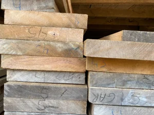 Pilhas de tábuas de madeira serrada, dispostas horizontalmente em duas colunas. As superfícies exibem marcações manuais em azul e preto, incluindo códigos como “5+”, “5h”, “6h”, “4S”, “5” e “6AK”. O cenário sugere organização em madeireira ou oficina, com identificação para medidas ou tipos de peças.