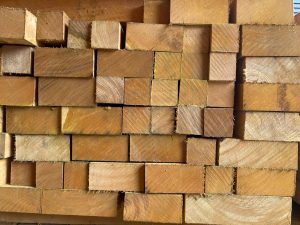 Pilhas de tábuas de madeira dispostas em formato compacto e horizontal, com extremidades retangulares mostrando marcas de serra e fibras soltas. As peças apresentam variações sutis de tamanho e coloração, indo do marrom claro ao médio, destacando textura natural e estado bruto, típicos de material para carpintaria e construção.
