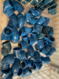 Coleção de conexões hidráulicas em PVC branco, formato Y, utilizadas para derivar tubulações em sistemas de encanamento. As peças estão empilhadas em recipiente de loja ou depósito. Algumas apresentam inscrições em relevo como “TIGRE” e “N35 50 50”, além de códigos de barras impressos. O ambiente sugere organização de materiais de construção e hidráulica para venda ou armazenamento.