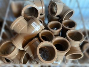 Conjunto de conexões plásticas marrons em formato de T, utilizadas em sistemas de esgoto, empilhadas em recipiente de loja ou depósito. As peças exibem marca Tigre e padrão NBR 5648, indicando conformidade técnica.