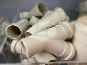 Coleção de conexões plásticas brancas em formato Y, utilizadas em sistemas hidráulicos para derivar tubulações. As peças, feitas em PVC, estão empilhadas em recipiente de loja ou depósito. Algumas apresentam inscrições em relevo como “TIGRE” e “N35 50 50”, além de códigos de barras impressos. O ambiente sugere organização de materiais de construção e encanamento para venda ou armazenamento.