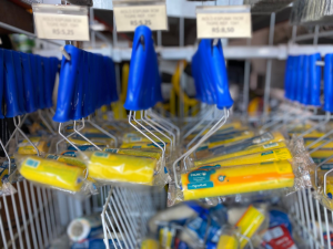 Estrutura metálica de exposição em loja, organizada com rolos de pintura de cabo plástico azul e espuma amarela, dispostos em fileiras suspensas. Alguns estão embalados em plástico transparente. Acima dos produtos, etiquetas de preço informam valores como “ROLO ESPUMA 9CM TIGRE MULTI 36U” por R$ 5,25 e R$ 5,50. O ambiente é bem iluminado e transmite organização típica de varejo de materiais de construção.