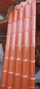 Grande chapa metálica laranja posicionada verticalmente em ambiente de depósito, com acabamento brilhante e padrão repetitivo de curvas que simulam telhas tradicionais. Ao fundo, há pilhas de materiais semelhantes e outros suprimentos de construção, reforçando o contexto de armazém de obras.