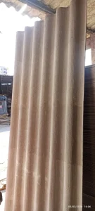 telha ondulada de material fibroso, em tom claro, posicionada verticalmente em ambiente externo ou semi-coberto. A superfície apresenta textura áspera típica de fibrocimento, usada em coberturas resistentes. Ao fundo, há pilhas de chapas semelhantes e tábuas de madeira, sugerindo depósito ou fábrica de materiais de construção.