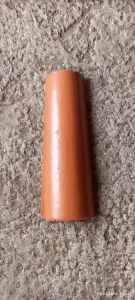 Peça cilíndrica de cerâmica em cor terracota, posicionada sobre superfície de concreto rugoso. O objeto apresenta formato afunilado, com uma extremidade mais larga que a outra, sugerindo função de conector ou adaptador em sistemas construtivos. O acabamento é liso e uniforme