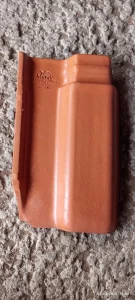 Telha cerâmica em tom terracota, modelo intertravado com perfil curvo e frisos moldados, posicionada sobre superfície de concreto rugoso. Na parte superior, inscrições em relevo indicam fabricante e dados técnicos: “Monier THE CONCRETE ROOF TILE ROMAN 1140mm x 720mm x 11mm 12.12.26P”. O objeto está isolado, em bom estado e destaca-se pelo acabamento uniforme.