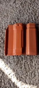 Telha cerâmica em tom terracota, posicionada sobre superfície de concreto rugoso. O design apresenta duas canaletas verticais paralelas e uma elevação central, característica de modelos intertravados usados em coberturas residenciais. A peça está íntegra, sem rachaduras ou marcas adicionais, destacando-se pelo acabamento uniforme e pela tonalidade avermelhada típica da cerâmica.