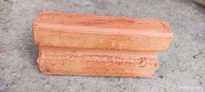 Tijolo cerâmico em tom vermelho, colocado sobre piso de concreto rugoso. O objeto apresenta perfil moldado com frisos e relevos, sugerindo uso ornamental em construções como cornijas ou acabamentos arquitetônicos. A superfície é áspera, com marcas visíveis de ferramentas e textura natural da argila.