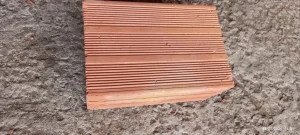 Peça quebrada de telha cerâmica em tom terracota, posicionada sobre superfície rugosa de concreto ou pedra. O fragmento apresenta frisos horizontais moldados, que podem servir para drenagem ou aderência, e está lascado em uma das extremidades, evidenciando desgaste ou dano. O acabamento é áspero e irregular, típico de materiais de construção