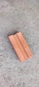 Tijolo cerâmico em tom vermelho-alaranjado, colocado sobre superfície de concreto rugoso com pequenas pedras incrustadas. A peça apresenta uma canaleta longitudinal moldada e está parcialmente danificada em uma das extremidades, com lasca visível. O acabamento é áspero, típico de tijolos de barro queimado usados em construção.
