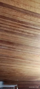 Forro de madeira com tábuas paralelas em tons naturais, luzes embutidas, parte de parede e prateleira metálica branca visíveis