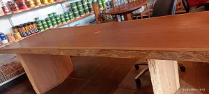 Mesa retangular de madeira com acabamento natural, tampo espesso e pés firmes, posicionada em área interna com piso de cerâmica. Ao fundo, prateleiras exibem latas e recipientes, possivelmente de tintas ou produtos domésticos. Outras mesas e cadeiras de madeira também estão visíveis, sugerindo espaço de exposição ou showroom. A iluminação é natural e uniforme