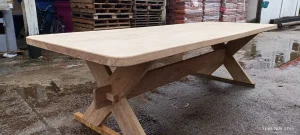 Mesa grande de madeira em tom natural, com tampo espesso de bordas arredondadas e base robusta em formato de X, reforçada por travessa central. A peça está posicionada em área externa sobre piso de concreto úmido. Ao fundo, aparecem pallets empilhados, bicicletas e materiais embalados, sugerindo ambiente de oficina ou depósito de madeira.