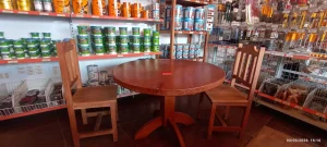 Mesa redonda de madeira em tom avermelhado, acompanhada por duas cadeiras com encosto em ripas verticais. Sobre o tampo, há um pequeno item vermelho, possivelmente etiqueta ou marcador. Ao fundo, prateleiras organizadas exibem latas de tinta e produtos diversos, enquanto na lateral direita ferramentas como chaves de fenda e alicates estão penduradas em suportes. Abaixo das prateleiras, cestos metálicos contêm mais materiais.