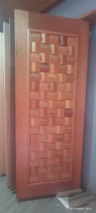 Versão expandida (≤300 caracteres): Porta de madeira posicionada verticalmente em ambiente interno, com painel central composto por blocos quadrados em grade, cada um com bordas chanfradas que criam efeito tridimensional semelhante a trama entrelaçada. A moldura externa é lisa e polida