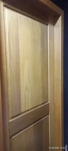 Close de porta de madeira em tom natural, composta por painel retangular central com veios verticais e detalhe horizontal próximo à base. A moldura externa acompanha a mesma textura e cor da porta. À direita, parede escura contrasta com o acabamento da madeira.