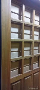 Estrutura de madeira clara em formato de porta ou divisória, composta por grade simétrica de retângulos e quadrados formados por ripas verticais e horizontais. Cada seção é preenchida por material translúcido semelhante a vidro fosco, permitindo passagem de luz com privacidade. O estilo remete a arquitetura tradicional japonesa