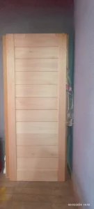 Porta de madeira em tom claro, composta por ripas horizontais uniformes e moldura vertical, posicionada de pé contra parede interna. À direita, vê-se parcialmente uma peça coberta por plástico, sugerindo área de armazenamento. O piso é revestido por azulejos de textura amadeirada e as paredes apresentam cores distintas: roxa à direita e tom escuro à esquerda.