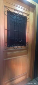 Porta de madeira em tom natural, composta por molduras retangulares e detalhes entalhados. No painel superior central, há grade de ferro forjado com curvas simétricas e barras verticais, unindo estética e segurança. O design ressalta o contraste entre a textura orgânica da madeira e o metal trabalhado.