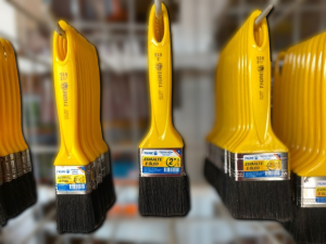 Expositor organizado em loja com pincéis Tigre de cabo amarelo e cerdas pretas, pendurados em fileiras. As embalagens trazem inscrições como “ESMALTE E ÓLEO” e códigos “519”. Os tamanhos visíveis variam entre “1 1/2”, “2” e “4” polegadas, destacados em etiquetas. A disposição transmite clareza e organização típica de ambiente varejista de materiais de pintura