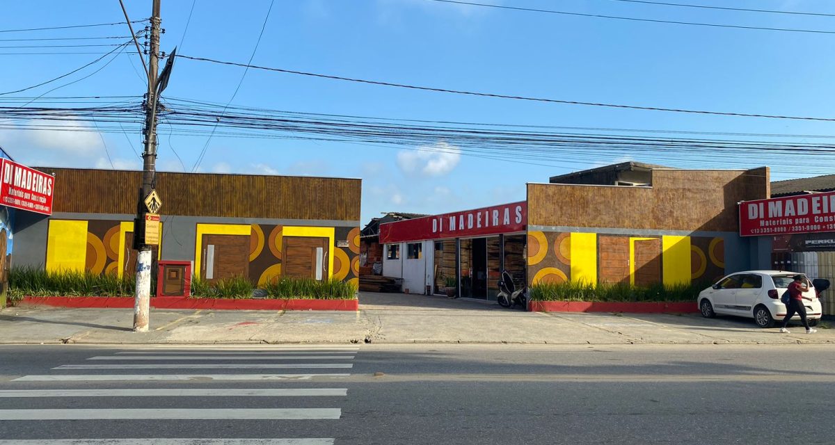 Fachada ampla da loja “DI MADEIRAS – Materiais para Construção”, com painéis amarelos e portas de madeira. Em frente, avenida com faixa de pedestres, poste com fiação aérea e céu azul. À direita, carro branco estacionado e uma pessoa caminhando na calçada